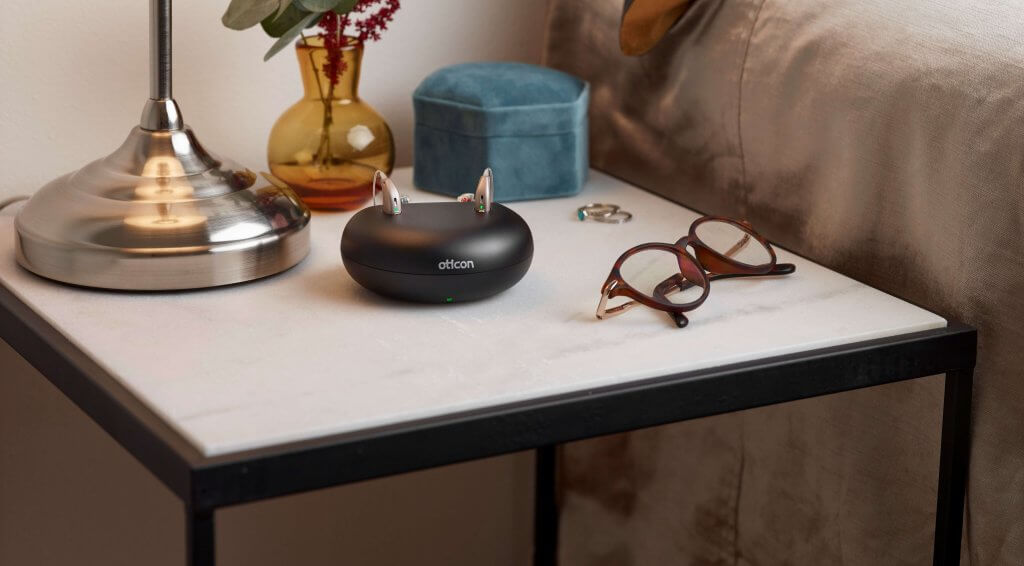 A pair of hearing aids Oticon Opn in a charging doc on a bedside table.