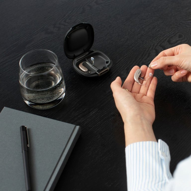 One Oticon Real hearing aid in the hand of a model, with the other hearing aid in it's charging case on a table. There is a a small glass of water, notepad and pen to the left hand side of the models hands, also on the table.