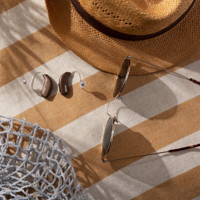 image of Oticon Real hearing aids, on a beach matt, with sunglasses and a sun hat.