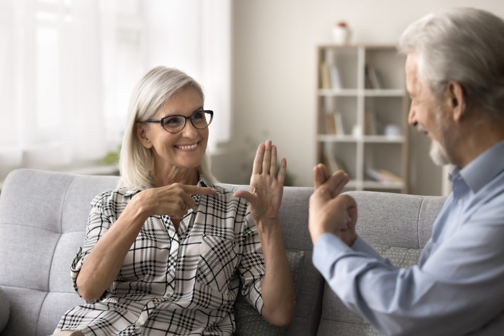 Smiling senior woman in glasses with a hearing disorder using hand gestures to communicate with her elderly husband.