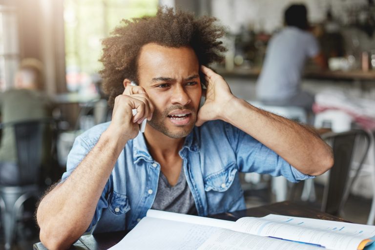 Frustrated man plugging one ear while trying to hear a phone conversation.