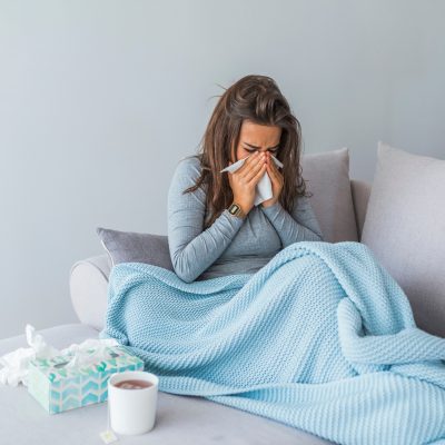 A woman looking unwell, sitting on a sofa and blowing her nose, with a cup of tea beside her and a blanket over her.