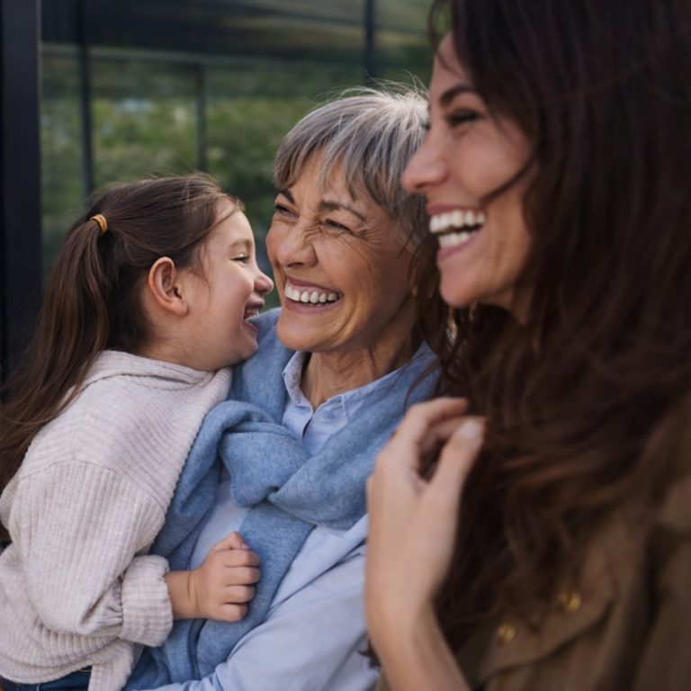 A grandmother, mother and daughter smiling together outdoors, looking into the distance.